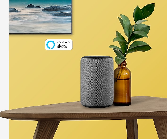 Corner of the SAMSUNG Q80R mounted on a yellow wall, on a wooden table with amazon alexa next to a bottle-potted plant