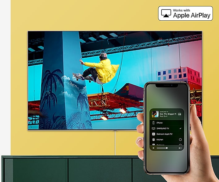 SAMSUNG Q80R Mounted on a Yellow Wall above a Green Cabinet. At the forefront is a hand holding a smartphone with AirPlay 2 up