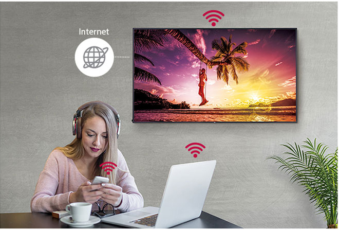 LG display showing a girl on a swing connected to a palm tree connected to a palm tree. A woman on her phone is using her smartphone in front of her open laptop with wifi signals showing all devices in the image are connected via the internet
