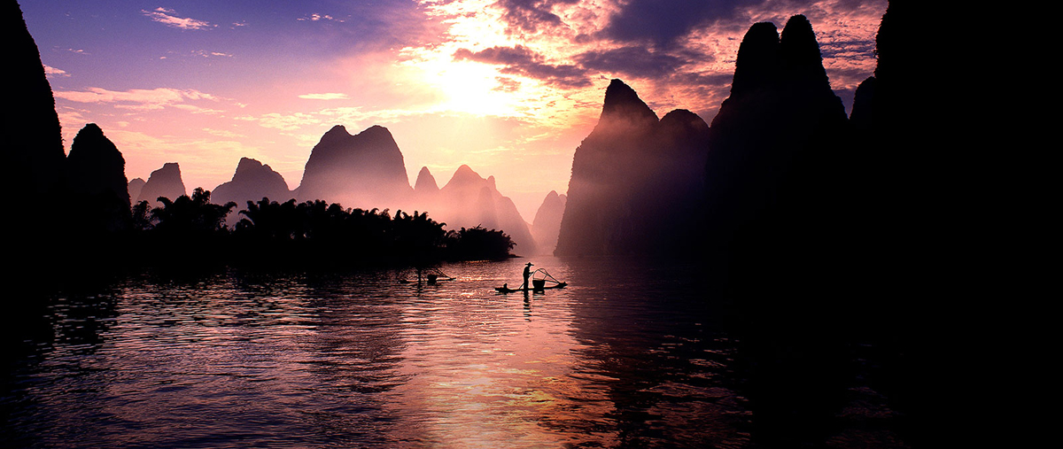 A scenery with two fishers on their own boats individually on a river surrounded by mountain ranges is in silhouette