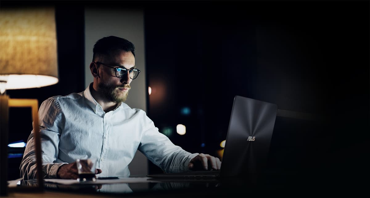 A man working at night looking and using his ASUS ZenBook Pro 15 UX550GE laptop indoors