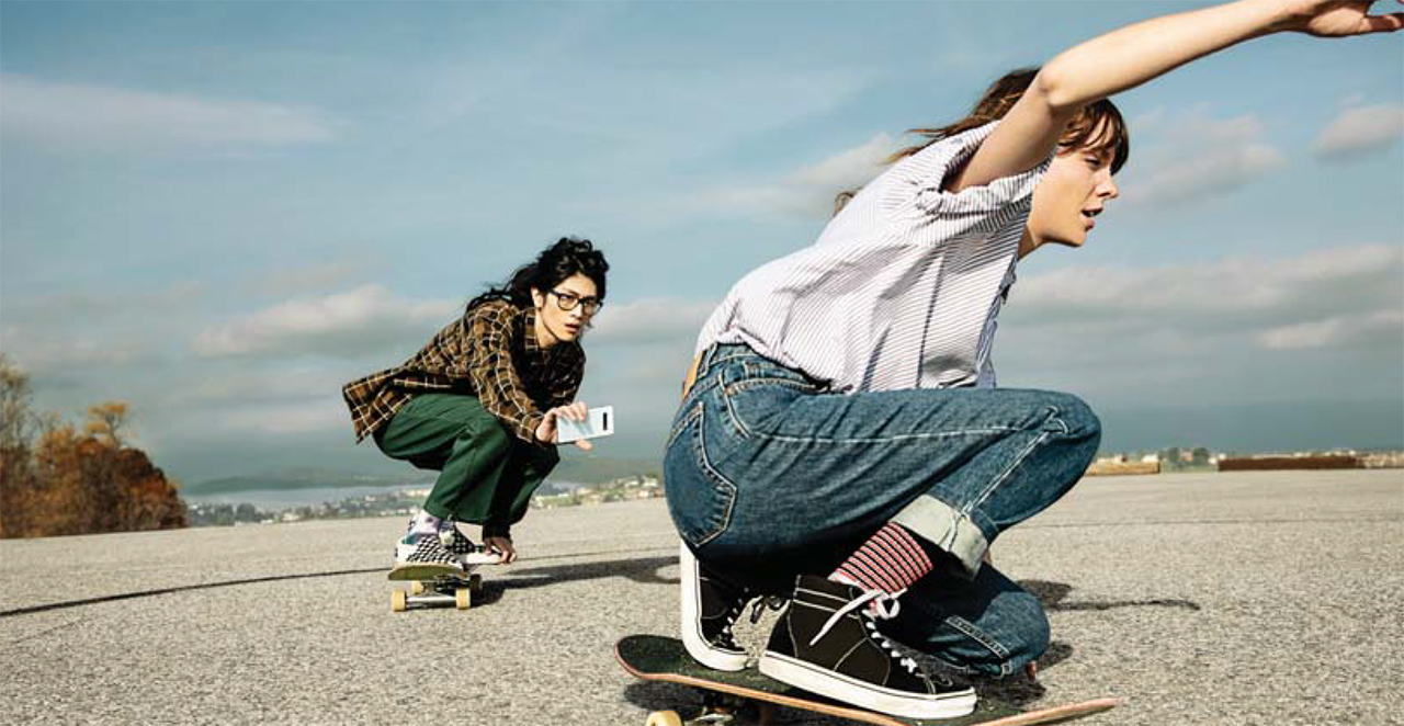 Two females skateboarding low with their knees bent. One is in front of the other and the female skater in the back is using the Samsung Galaxy S10 in her left hand to film the female skater in front of her