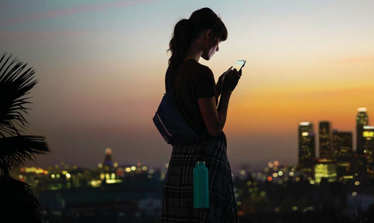 A woman with her hiking backpack and attached water flash looking down on her Samsung Galaxy S10 between both her hands in front of the Los Angeles skyline at twilight