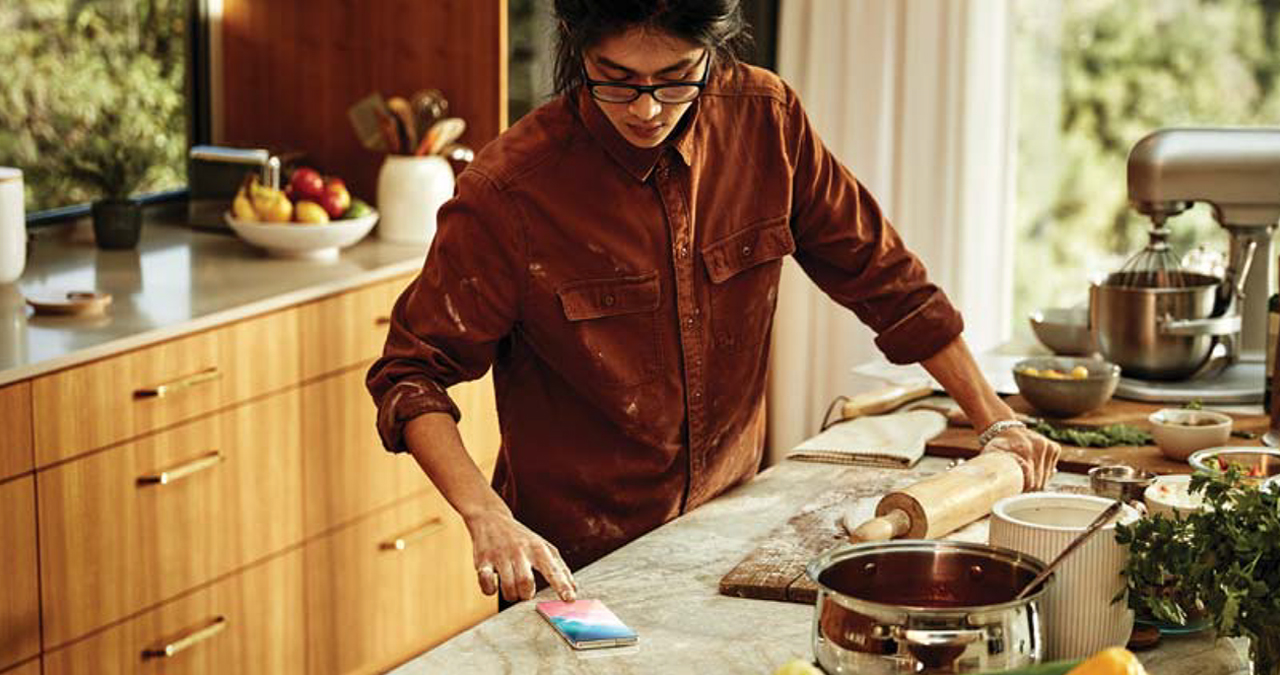 A man in the kitchen using a roller on dough, taking a break to unlock his Samsung Galaxy S10 with his right index finger