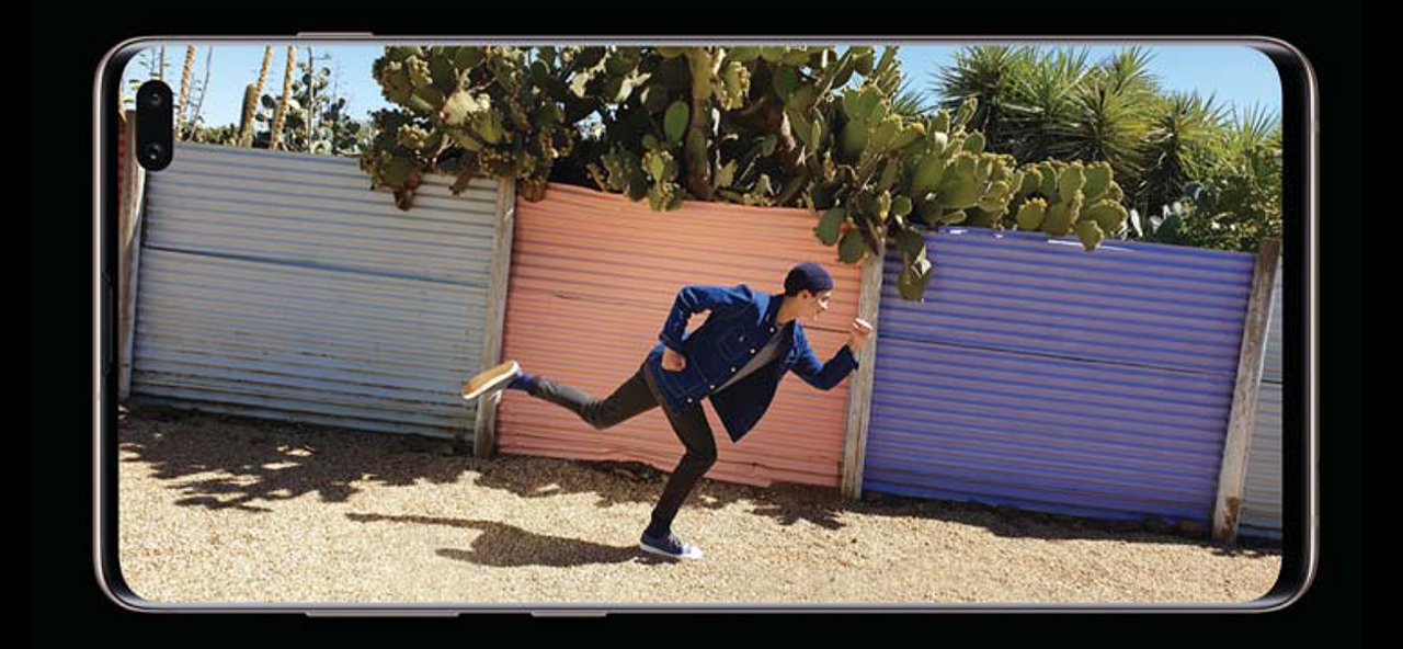A Samsung Galaxy S10 sitting horizontally on its side showing a young man posing as if he were on the go in front of three colored sheet-metal fences and overgrowing cacti