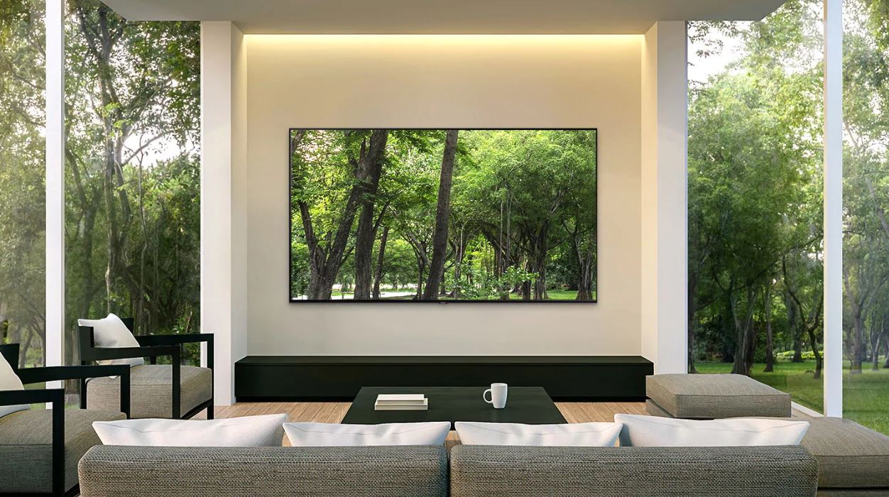 A Samsung Q70R display mounted on a wall in a living room with high windows and trees outdoors