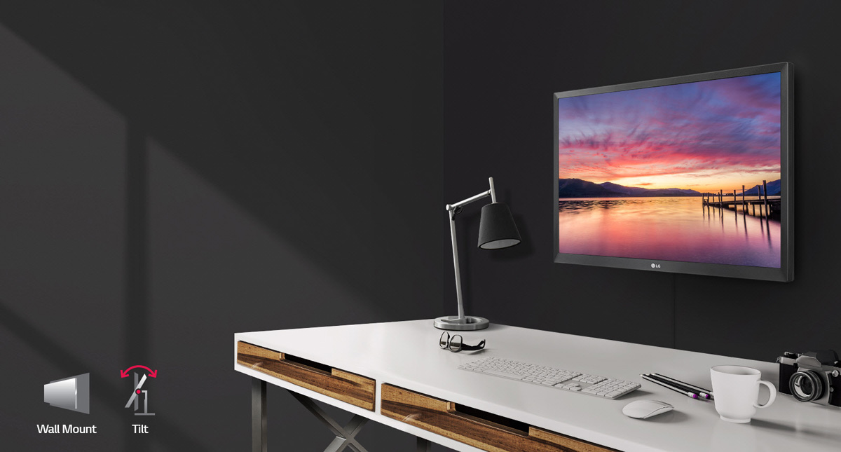 LG 24BK430H-B Monitor Wall Mounted Angled to the Left in Front of a Desk that has a lamp, glasses, keyboard, mouse, pencils, coffee mug and camera. At the bottom left of this image is a wall mount and tilt icon