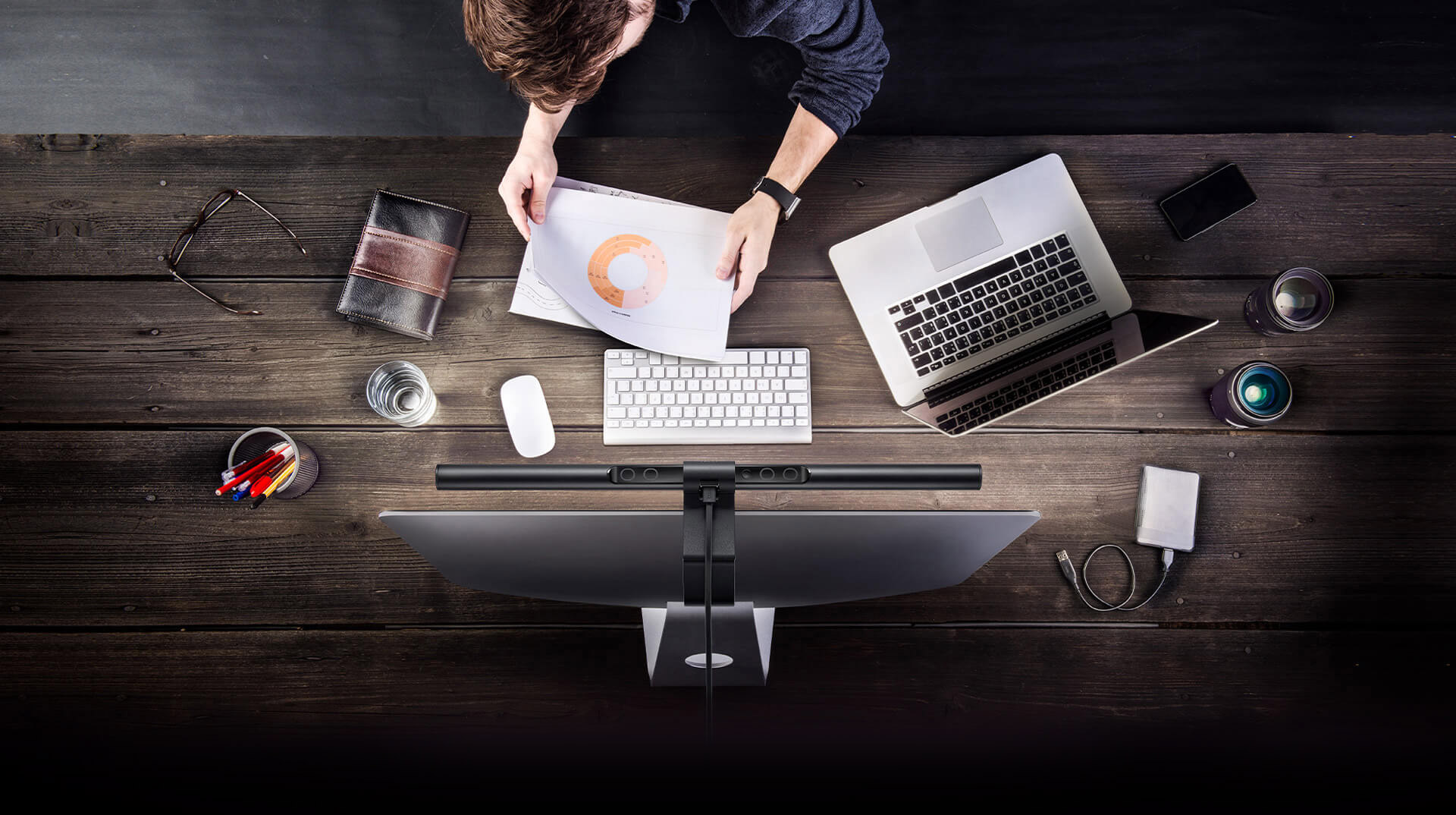 overhead view of the BenQ ScreenBar Plus on top of an imac with a man working on a desk that has a cup of pens, glasses, leather-bound notebook, cup of water, mouse, keyboard, open laptop, smartphone, camera lenses and external hard drive with cable