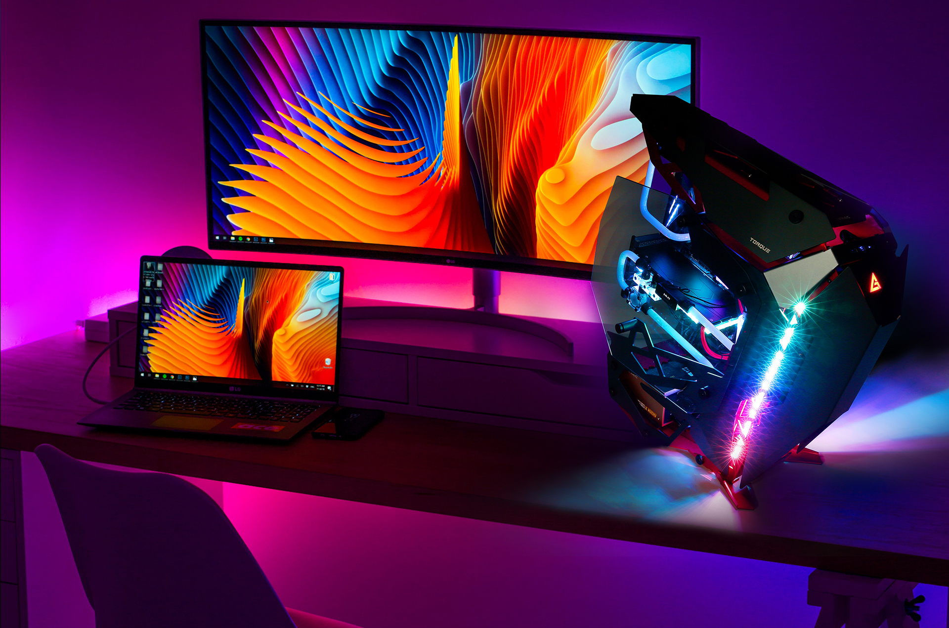 Antec TORQUE on a desk with a laptop plugged into a wide-view monitor, both screens show a blue and orange graphic that looks like an electric canyon