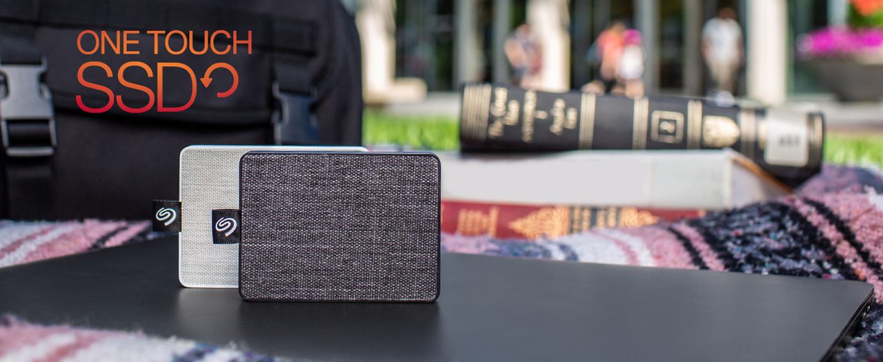 A One Touch in black and a One Touch in White in standing position placed on a laptop, in front of a bag and some books. The background is some students on the campus