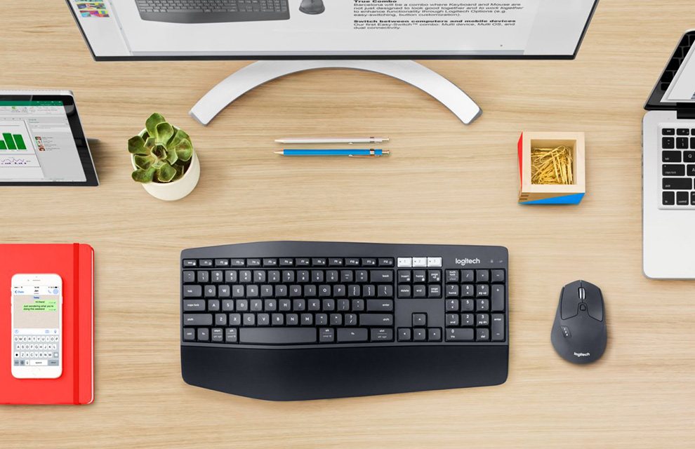 Overhead view of the Logitech MK850 Performance Wireless Keyboard and Mouse Combo on a wooden desktop in front of a monitor and box of yellow paperclips