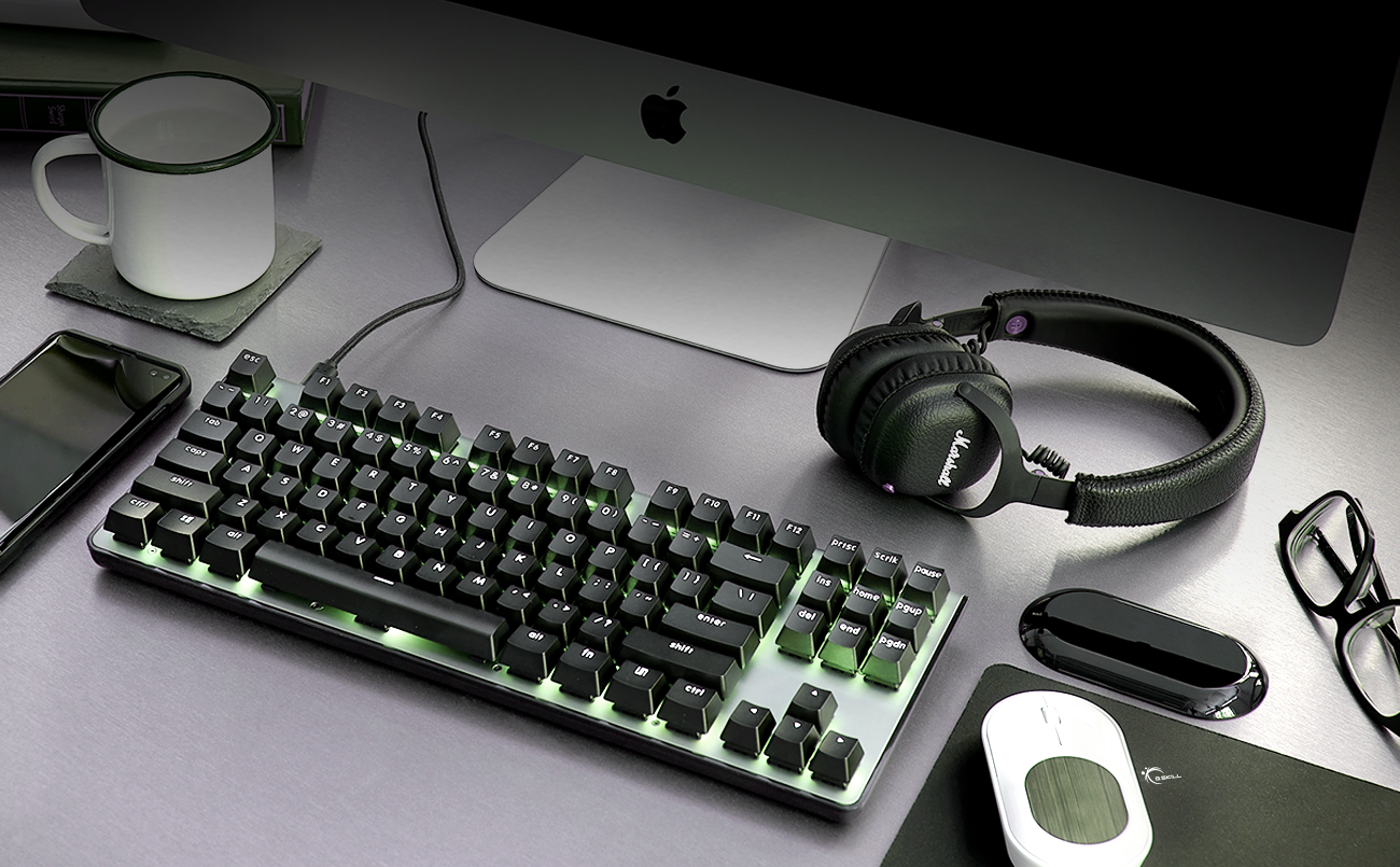 a black keyboard with a backlighting, a headphone, a mouse, a glasses, a cup of water, a phone and a computer