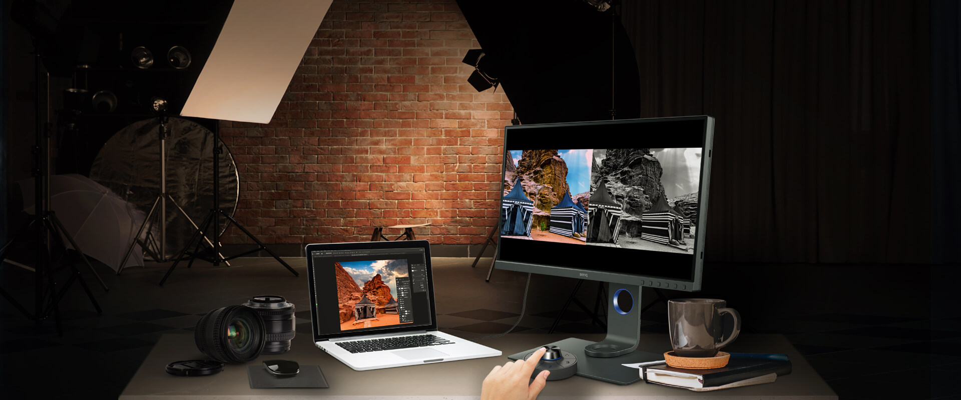 BenQ SW270C monitor on a desk angled to the left along with a DSLR camera and accessories, laptop with photo-editing software on screen, a control button, books and a coffee mug. Behind the desk is a brick wall and photo lighting equipment