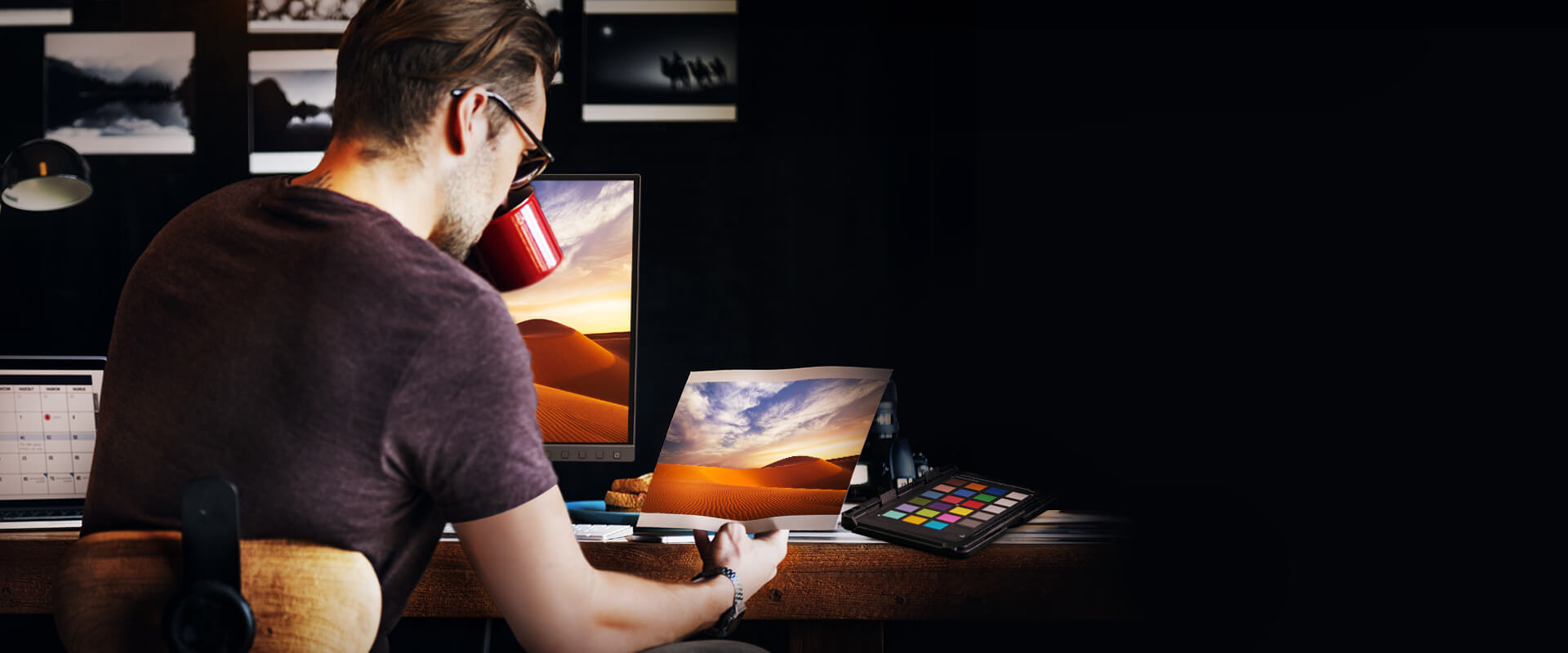 A Male Designer at his wooden desk with the BenQ SW270C monitor, a tablet he's holding, another tablet with the home screen open and headphones behind him on his wooden chair