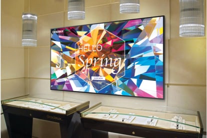 Samsung Display Mounted Above a couple of arched glass-covered tables with jewelry inside. The Samsung display is showing a spring multi-colored, geometric pattern ad on screen