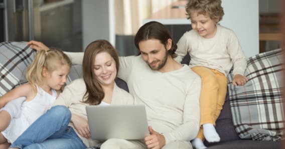 A family of four gathererd around a laptop while on their living room couch